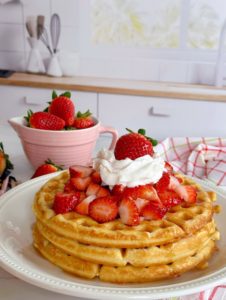 Stack of three Vanilla Waffles topped with fresh Strawberries and whipped cream and a small bowl of strawberries behind.