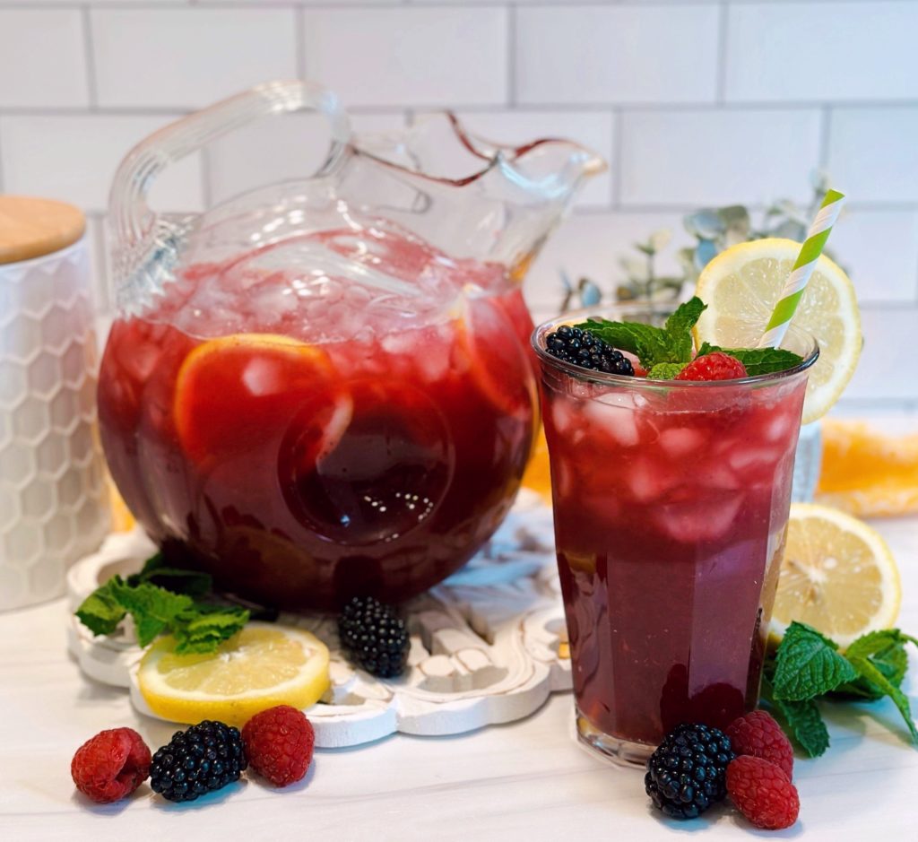 Triple Berry Lemonade in a beautiful crystal pitcher and a tall glass poured with lemon wedges, mint, and fresh berries.