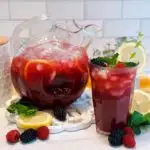 Triple Berry Lemonade in a beautiful crystal pitcher and a tall glass poured with lemon wedges, mint, and fresh berries.