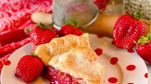 Big slice of Strawberry Rhubarb Pie on a fun polka dot plate with fresh strawberries surrounding it and a pretty background.