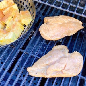 Two chicken marinated chicken breast on the BBQ grill with a grill basket full of pineapple topped with brown sugar and cinnamon.