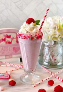 White Chocolate Raspberry Milk Shake in an old fashioned milk shake glass rimmed with valentine sprinkles, topped off with a cloud of whipped cream and a fresh raspberry and fun striped straw.