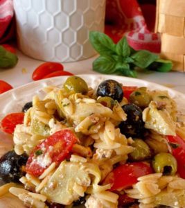 Muffuletta Olive Pasta Salad close up on a white plate with ingredients scattered around it.