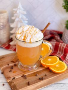 Glass of Old Fashioned Spiced Apple Cider on a cutting board with orange slices surrounding it. Topped with Whipped Cream and a caramel drizzle.