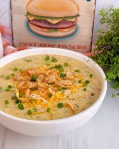 Creamy Cheeseburger Soup in a white bowl on a marble counter top with a photo of burger in the background. Topped with green onions and fried crunchy onions.