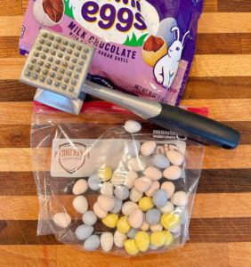 Candy eggs in a ziploc bag with a meat mallet on a wooden cutting board.