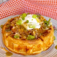 Cornbread waffle stuffed with chili and cheese and topped with sour cream and more cheese on a dinner plate.