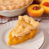 Fresh baked peach pie slice on a dessert dish with fresh peaches and whole pie in the background.