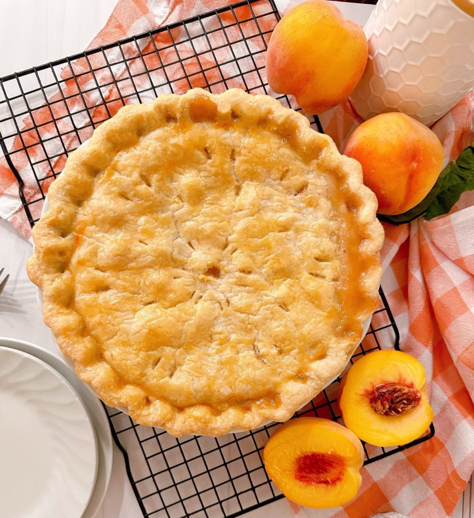 Fresh baked peach pie on a cooling rack surrounded with fresh peaches.