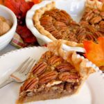 Slice of the Best Pecan Pie Recipe on a white dessert plate with a fork and the whole pie in the background.