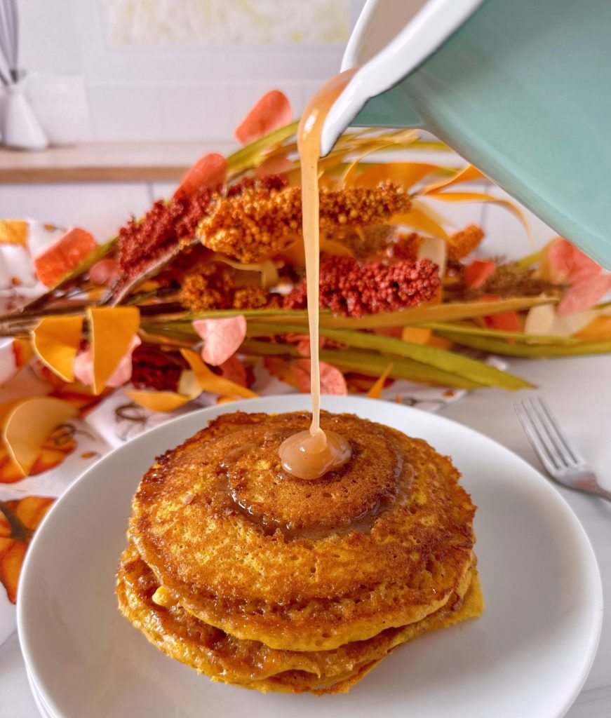 Pouring cinnamon bun syrup over the top of a stack of pumpkin cinnamon pancakes.