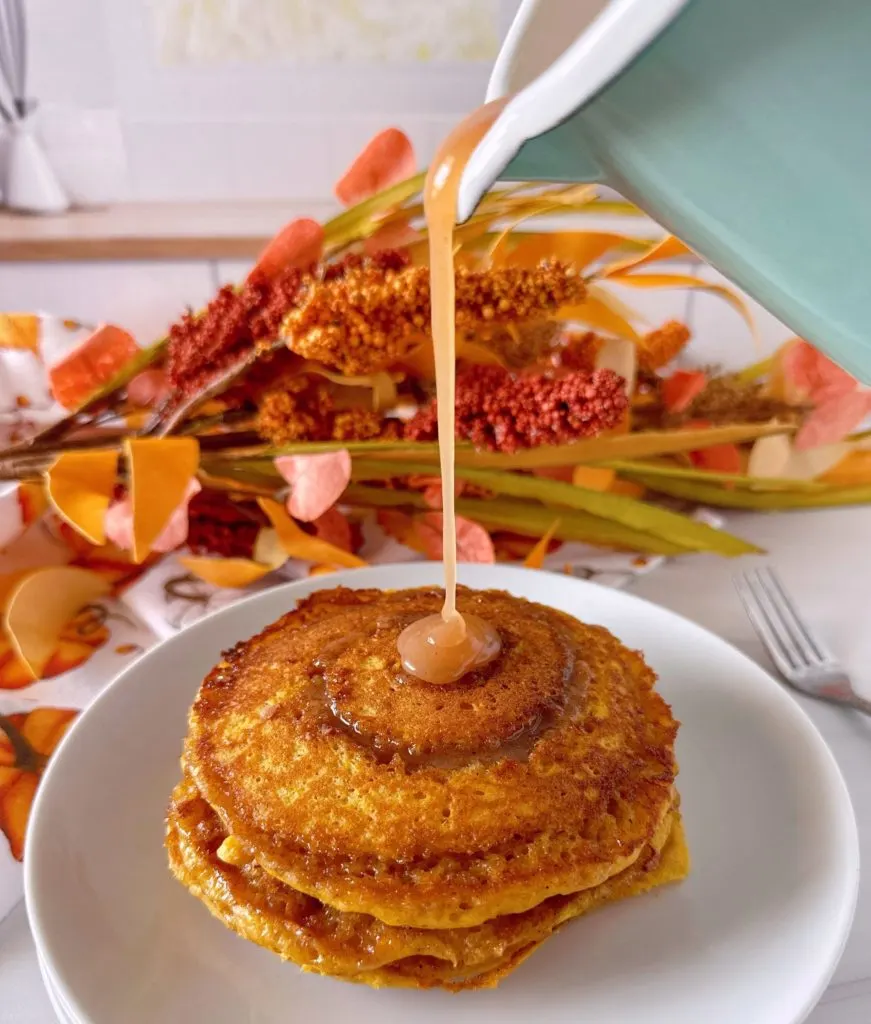 Pouring cinnamon bun syrup over the top of a stack of pumpkin cinnamon pancakes.